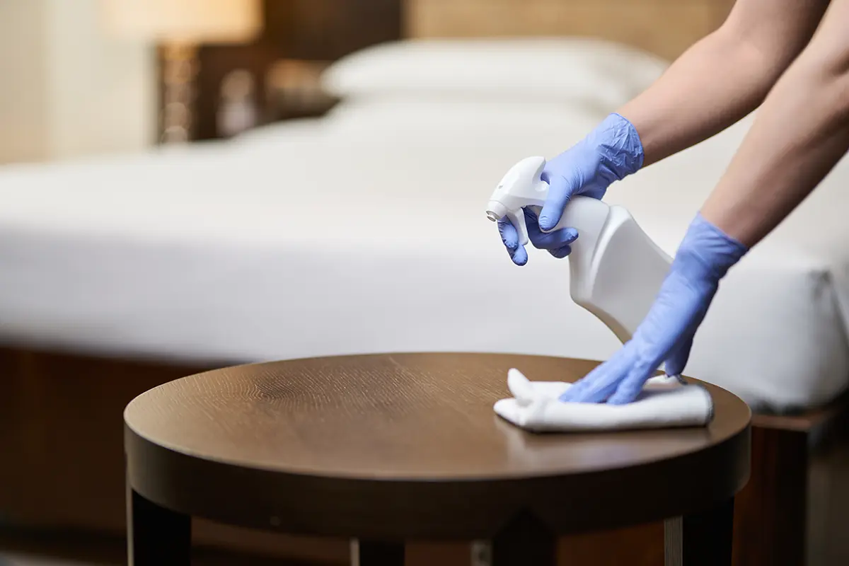 A housemaid wiping down the bedside table with cleaning spray in an upscale bedroom.